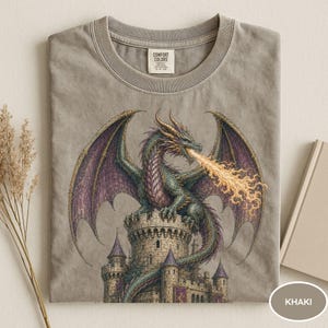 May include: A khaki-colored t-shirt featuring a graphic print of a green and purple dragon perched atop a stone castle tower, breathing a stream of orange fire. The shirt has a crew neckline and a Comfort Colors brand label at the inner collar.