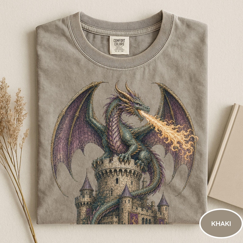 May include: A khaki-colored t-shirt featuring a graphic print of a green and purple dragon perched atop a stone castle tower, breathing a stream of orange fire. The shirt has a crew neckline and a Comfort Colors brand label at the inner collar.