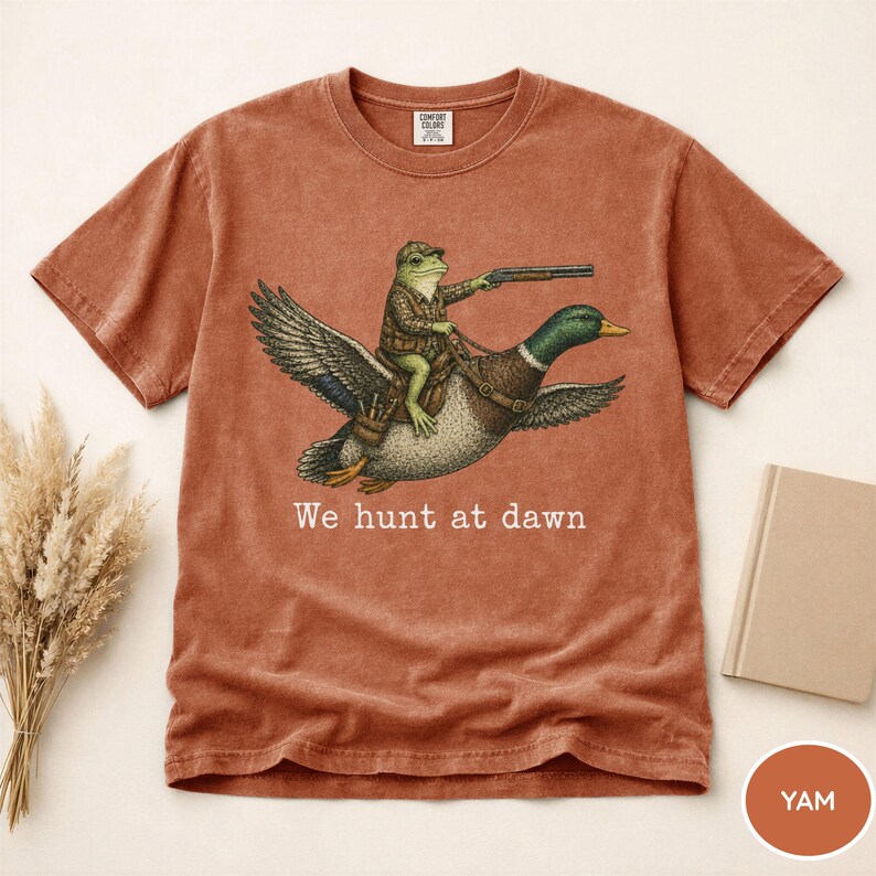 Puede incluir: Camiseta de color terracota con una ilustraci&oacute;n caprichosa de una rana montada en un pato y sosteniendo un rifle. El texto "We hunt at dawn" est&aacute; impreso debajo. Cuello redondo, mangas cortas.