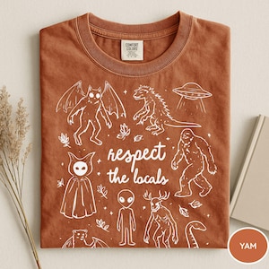 May include: A rust-colored Comfort Colors t-shirt featuring white illustrations of various cryptids and a UFO. The phrase "respect the locals" is printed in the center. The shirt is folded neatly.