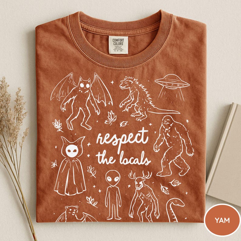 May include: A rust-colored Comfort Colors t-shirt featuring white illustrations of various cryptids and a UFO. The phrase "respect the locals" is printed in the center. The shirt is folded neatly.