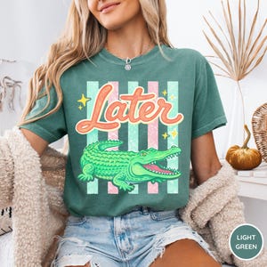 May include: A light green t-shirt featuring a cartoon alligator and the word "Later" in a playful font. The design includes pink and light green stripes and yellow stars. The shirt is a comfortable, casual style.