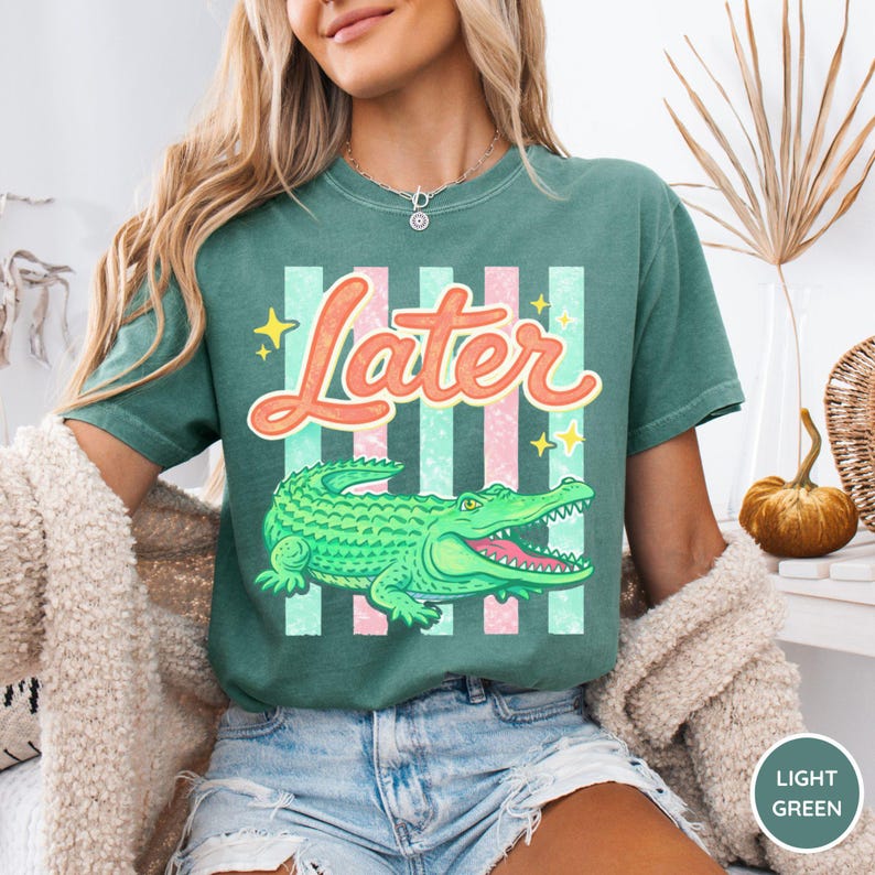 May include: A light green t-shirt featuring a cartoon alligator and the word "Later" in a playful font. The design includes pink and light green stripes and yellow stars. The shirt is a comfortable, casual style.