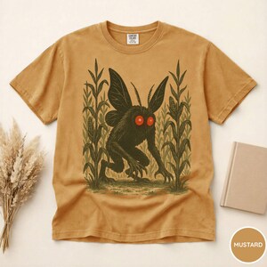 K&ouml;nnte beinhalten: Senffarbenes T-Shirt mit einer Grafik eines kryptischen Mottenwesens mit roten Augen, das in einem Maisfeld steht. Das Shirt hat einen Rundhalsausschnitt und kurze &Auml;rmel. Der Text "COMFORT COLORS" ist auf dem oberen R&uuml;cken des Shirts zu sehen.