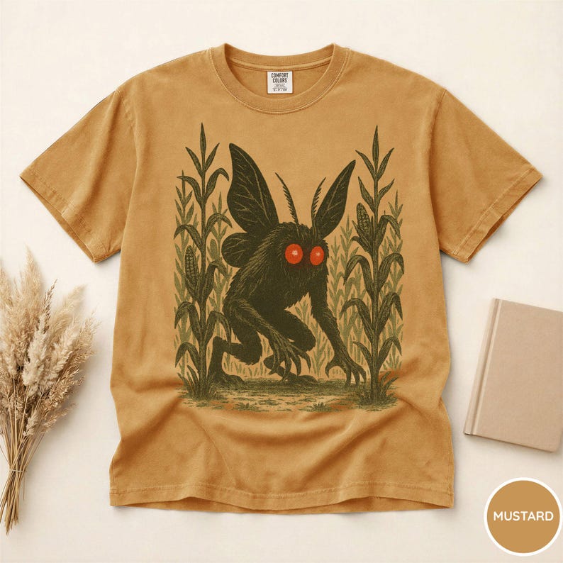 K&ouml;nnte beinhalten: Senffarbenes T-Shirt mit einer Grafik eines kryptischen Mottenwesens mit roten Augen, das in einem Maisfeld steht. Das Shirt hat einen Rundhalsausschnitt und kurze &Auml;rmel. Der Text "COMFORT COLORS" ist auf dem oberen R&uuml;cken des Shirts zu sehen.