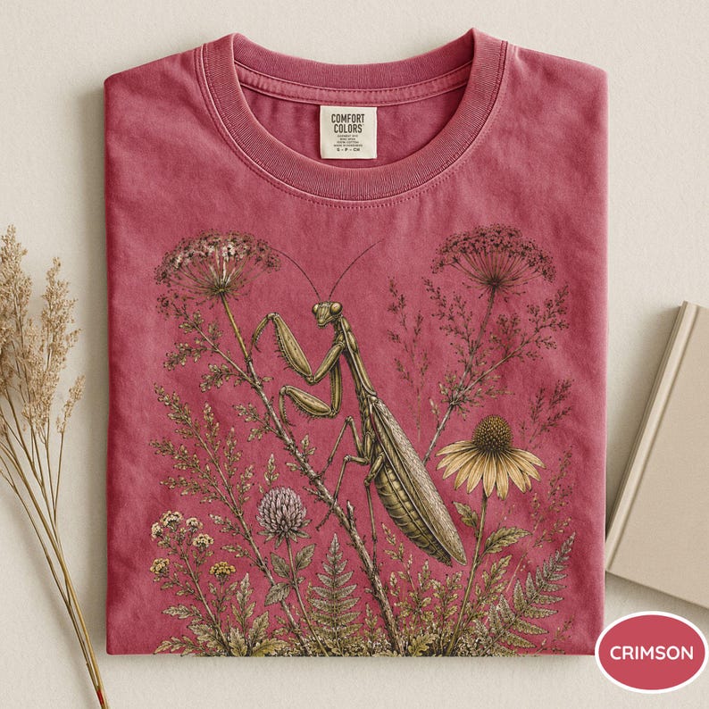May include: Crimson-colored t-shirt featuring a detailed illustration of a praying mantis amidst wildflowers and foliage. The design includes a variety of plants and a yellow flower. The shirt has a crew neck and a tag that says "Comfort Colors".