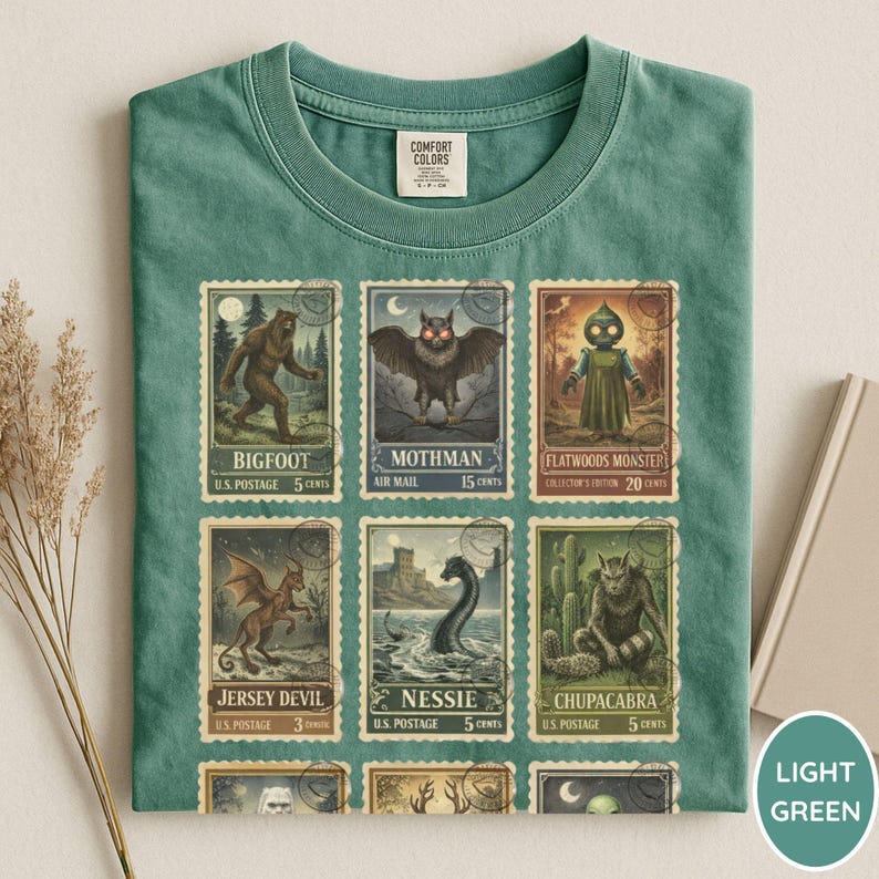 May include: Light green t-shirt featuring a collection of vintage-style postage stamps with cryptid illustrations, including Bigfoot, Mothman, and Nessie. The shirt has a crew neck and a Comfort Colors tag.