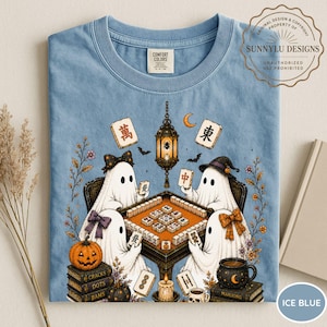 May include: An ice blue t-shirt featuring a graphic of four ghosts playing mahjong at a table. The design includes a hanging lantern, a pumpkin, stacks of books labeled Cracks, Dots, and Bams, and various mahjong tiles. The shirt has a Comfort Colors label.