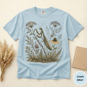 May include: Light blue t-shirt featuring a detailed illustration of a praying mantis amidst wildflowers and foliage. The design includes a variety of plants and a yellow flower. The shirt has a crew neck and short sleeves. The text "CHAM BRAY" is in a circle.