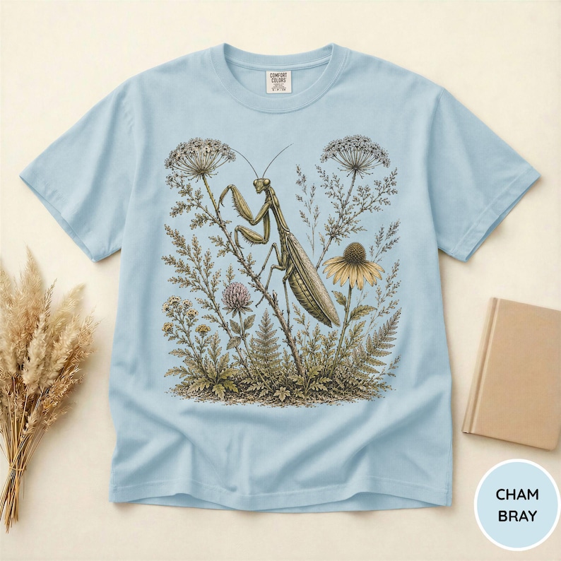 May include: Light blue t-shirt featuring a detailed illustration of a praying mantis amidst wildflowers and foliage. The design includes a variety of plants and a yellow flower. The shirt has a crew neck and short sleeves. The text "CHAM BRAY" is in a circle.