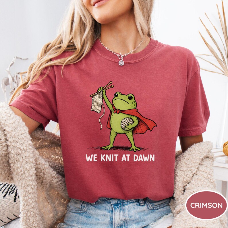 May include: Crimson-colored t-shirt featuring a cartoon frog wearing a red cape and holding knitting needles and yarn. The text "WE KNIT AT DAWN" is printed below the frog. The shirt is a comfortable, casual style.