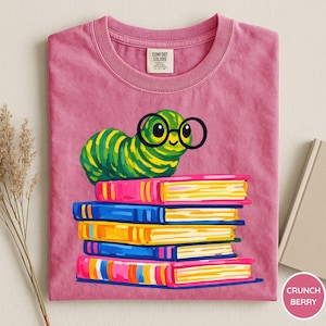 Pu&ograve; includere: Una t-shirt rosa con la grafica di un bruco a strisce verdi e gialle che indossa occhiali tondi neri, appoggiato su una pila di cinque libri colorati. La maglietta presenta un'etichetta del marchio Comfort Colors all'interno del colletto.