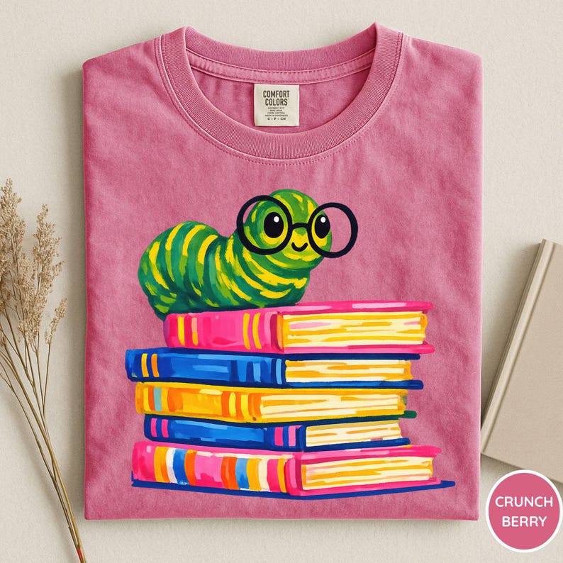 Pu&ograve; includere: Una t-shirt rosa con la grafica di un bruco a strisce verdi e gialle che indossa occhiali tondi neri, appoggiato su una pila di cinque libri colorati. La maglietta presenta un'etichetta del marchio Comfort Colors all'interno del colletto.