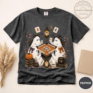 May include: A charcoal gray short-sleeve t-shirt featuring a graphic of four white ghosts playing mahjong around a table. The design includes a hanging lantern, a pumpkin, stacks of books, a skull mug, and floating mahjong tiles with Chinese characters.