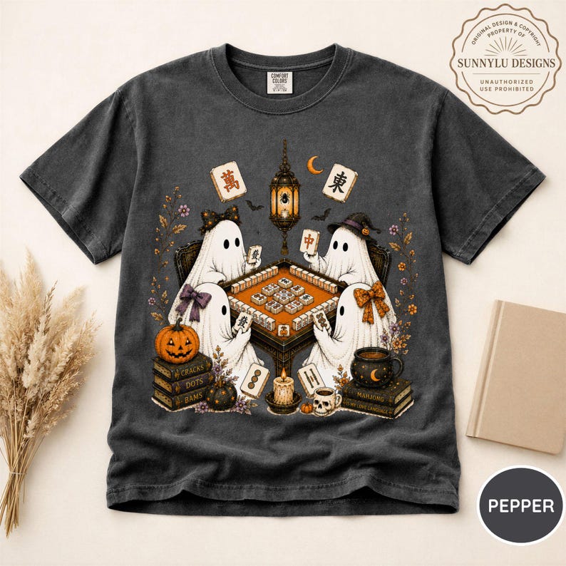 May include: A charcoal gray short-sleeve t-shirt featuring a graphic of four white ghosts playing mahjong around a table. The design includes a hanging lantern, a pumpkin, stacks of books, a skull mug, and floating mahjong tiles with Chinese characters.