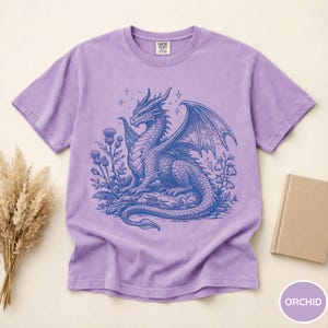 Peut inclure: T-shirt couleur orchid&eacute;e avec un motif de dragon bleu d&eacute;taill&eacute;, ailes d&eacute;ploy&eacute;es, entour&eacute; de fleurs et d'&eacute;toiles. Col rond, en mati&egrave;re douce.