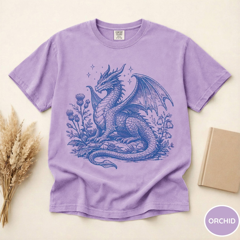 Peut inclure: T-shirt couleur orchid&eacute;e avec un motif de dragon bleu d&eacute;taill&eacute;, ailes d&eacute;ploy&eacute;es, entour&eacute; de fleurs et d'&eacute;toiles. Col rond, en mati&egrave;re douce.