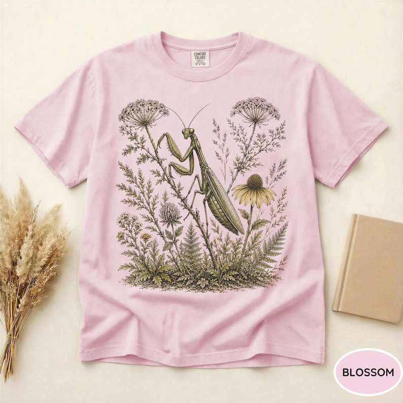 May include: A light pink t-shirt featuring a detailed illustration of a praying mantis amidst wildflowers and foliage. The design is in shades of green, brown, and yellow. The shirt has a round neckline and short sleeves. The word "BLOSSOM" is in a pink circle.