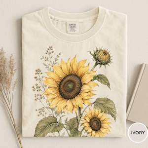 May include: Ivory-colored t-shirt featuring a watercolor illustration of sunflowers. The design includes a large, open sunflower with yellow petals and a dark brown center, along with smaller sunflowers and green leaves. The shirt has a crew neck and short sleeves.