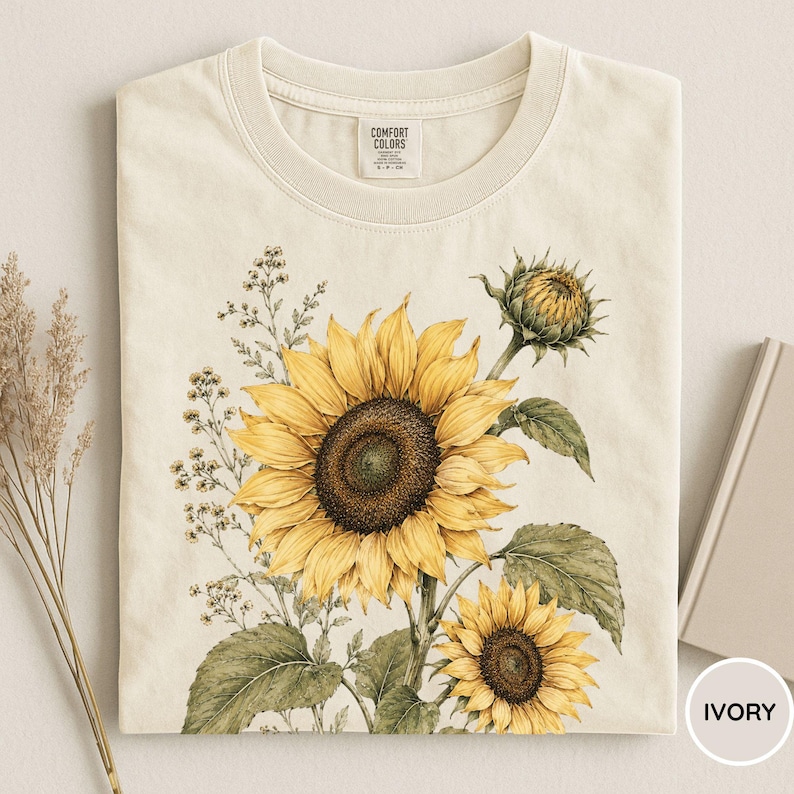 May include: Ivory-colored t-shirt featuring a watercolor illustration of sunflowers. The design includes a large, open sunflower with yellow petals and a dark brown center, along with smaller sunflowers and green leaves. The shirt has a crew neck and short sleeves.