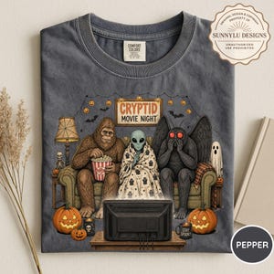 May include: A charcoal gray t-shirt featuring a graphic of Bigfoot, an alien, and Mothman sitting on a couch watching a television. The text reads "CRYPTID MOVIE NIGHT". The design includes pumpkins, a lamp, and a ghost, all set against a dark background.