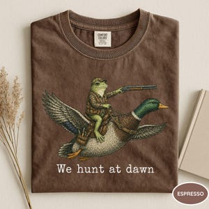Puede incluir: Camiseta color espresso con una ilustraci&oacute;n humor&iacute;stica de una rana con atuendo de caza montada en un pato, apuntando con una escopeta. El texto "We hunt at dawn" est&aacute; impreso debajo. Cuello redondo cl&aacute;sico.
