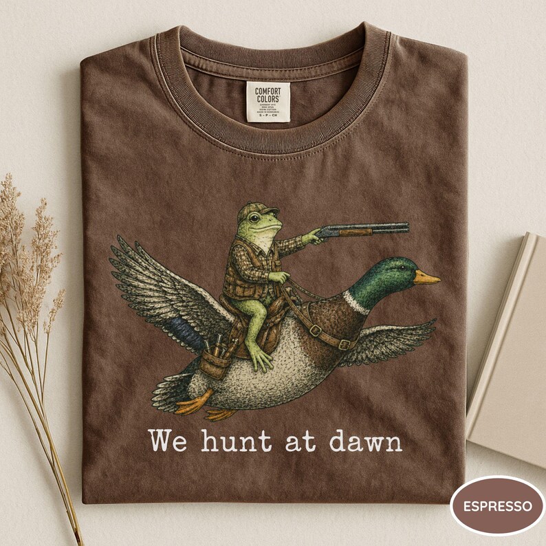 Puede incluir: Camiseta color espresso con una ilustraci&oacute;n humor&iacute;stica de una rana con atuendo de caza montada en un pato, apuntando con una escopeta. El texto "We hunt at dawn" est&aacute; impreso debajo. Cuello redondo cl&aacute;sico.