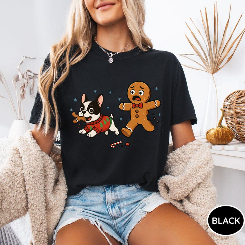 May include: Black t-shirt featuring a cartoon dog in a Christmas sweater chasing a gingerbread man. The dog is holding a gingerbread cookie, and the gingerbread man has a worried expression. The shirt has a round neck.