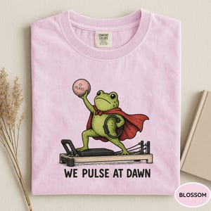 May include: A light pink t-shirt featuring a graphic of a green frog wearing a red cape, standing on a pilates reformer machine. The frog holds a pink ball labeled Pilates and a black ring. The text below reads WE PULSE AT DAWN.