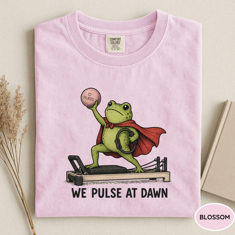 May include: A light pink t-shirt featuring a graphic of a green frog wearing a red cape, standing on a pilates reformer machine. The frog holds a pink ball labeled Pilates and a black ring. The text below reads WE PULSE AT DAWN.