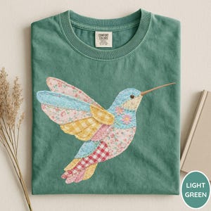 May include: Light green t-shirt featuring a colorful hummingbird applique. The hummingbird has patchwork wings with floral, gingham, and solid patterns in pink, yellow, and blue. The shirt has a crew neck and a tag on the collar.