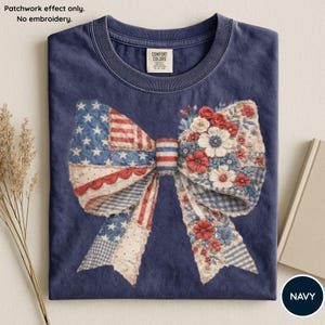 Op de afbeelding: Marineblauw T-shirt met een groot strikdesign. De strik heeft aan de ene kant een Amerikaans vlagpatroon en aan de andere kant bloemaccenten. Het T-shirt heeft een ronde hals en een label met de tekst "Comfort Colors".