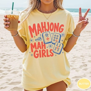May include: A pale yellow short-sleeve t-shirt featuring red text that reads "MAHJONG with MAH GIRLS" alongside blue illustrations of three mahjong tiles and small flower accents. The shirt has a crew neckline and a relaxed, loose-fitting cut.
