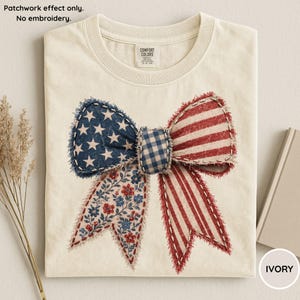 Pode incluir: Camiseta de cor marfim com um design de laço de patchwork. O laço incorpora padrões patrióticos: azul com estrelas brancas, listras vermelhas e brancas e estampas florais. A camiseta tem gola redonda e uma etiqueta Comfort Colors.