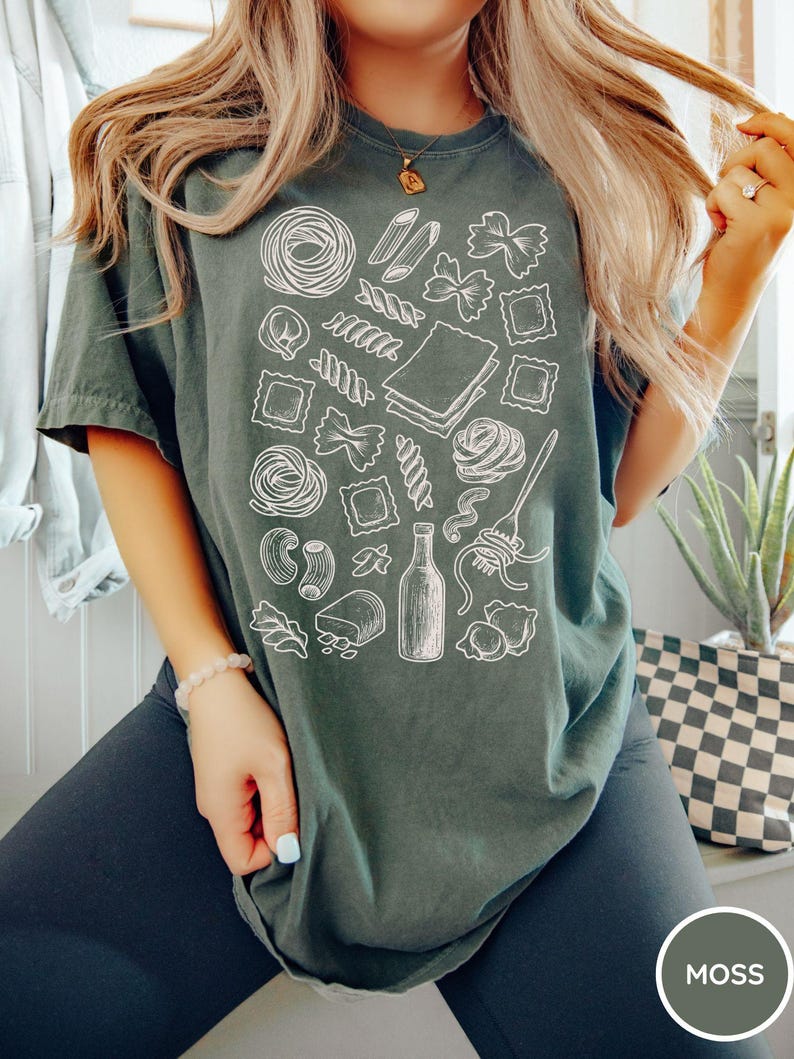 Puede incluir: Una camiseta de manga corta color verde musgo con un estampado gr&aacute;fico de l&iacute;neas blancas de varias formas de pasta, incluyendo farfalle, raviolis y penne, junto a una botella y un tenedor enrollando espaguetis, en un patr&oacute;n disperso.