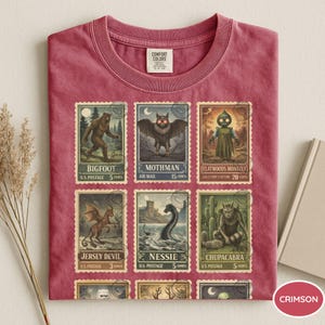 May include: Crimson-colored t-shirt featuring a collection of vintage-style postage stamps. Each stamp depicts a different cryptid, including Bigfoot, Mothman, and the Jersey Devil. The shirt has a crew neck and short sleeves.