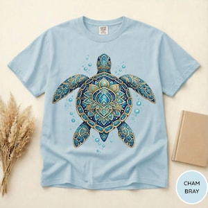May include: A light blue t-shirt featuring a central graphic of a sea turtle with an intricate, mandala-inspired shell pattern in shades of blue, teal, and gold, surrounded by small bubbles. The inner neck label displays the text Comfort Colors.