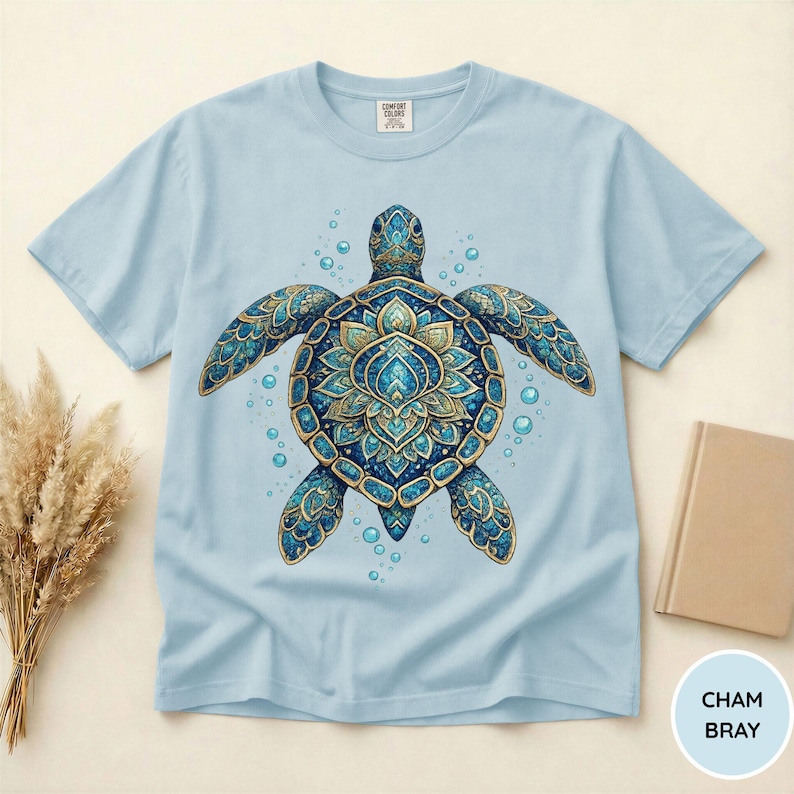 May include: A light blue t-shirt featuring a central graphic of a sea turtle with an intricate, mandala-inspired shell pattern in shades of blue, teal, and gold, surrounded by small bubbles. The inner neck label displays the text Comfort Colors.