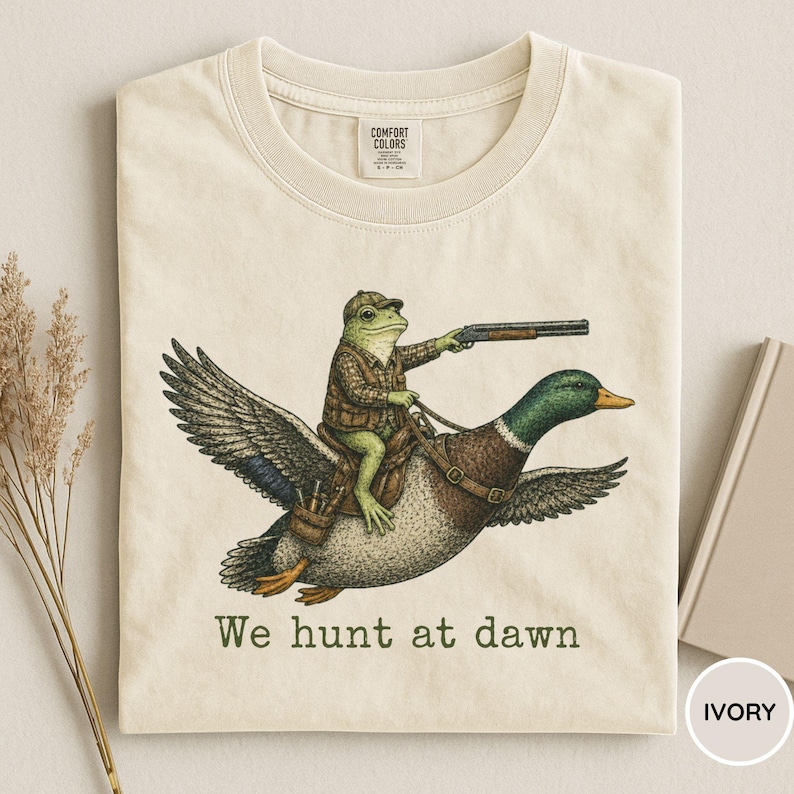 Puede incluir: Camiseta de color marfil con una ilustraci&oacute;n humor&iacute;stica de una rana con ropa de caza montada en un pato y sosteniendo una escopeta. El texto "We hunt at dawn" est&aacute; impreso debajo. La camiseta es de material suave.