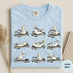 K&ouml;nnte beinhalten: Hellblaues T-Shirt mit einem sich wiederholenden Muster von Cartoon-Enten, die Pilates-Reformer benutzen. Die Enten befinden sich in verschiedenen &Uuml;bungspositionen. Der Text "CHAM BRAY" befindet sich in einem Kreis unten rechts. Das T-Shirt hat ein Comfort Colors-Etikett.