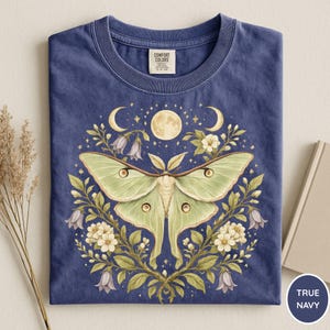 May include: True navy blue t-shirt featuring a detailed illustration of a luna moth with a full moon, crescent moons, and floral elements. The moth is light green with brown accents, surrounded by white flowers and purple bellflowers.