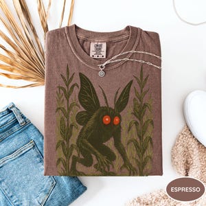 K&ouml;nnte beinhalten: Espressofarbenes T-Shirt mit einem Aufdruck einer Kreatur mit Fl&uuml;geln und roten Augen, in einem Maisfeld. Das Shirt hat einen Rundhalsausschnitt und ein Comfort Colors-Etikett. Eine silberne Halskette mit Sonnenanh&auml;nger liegt auf dem Shirt.