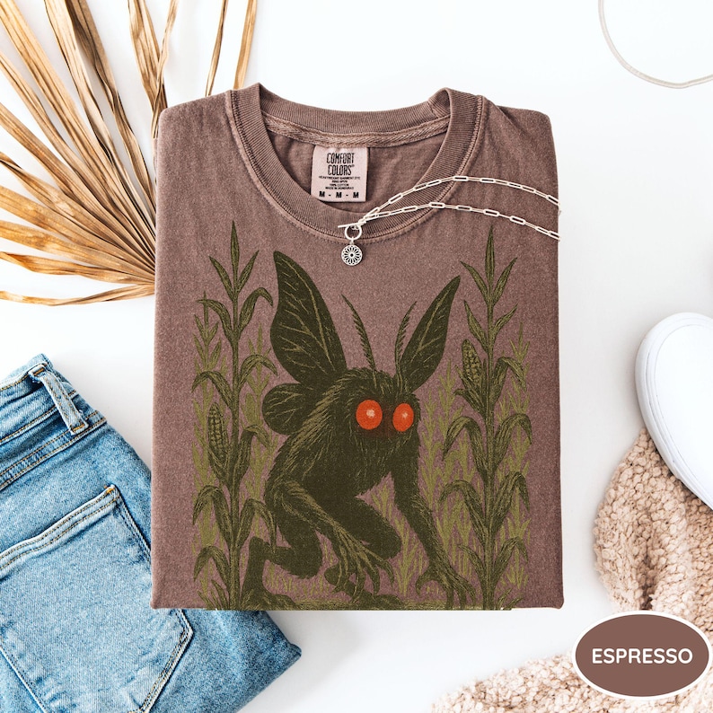 K&ouml;nnte beinhalten: Espressofarbenes T-Shirt mit einem Aufdruck einer Kreatur mit Fl&uuml;geln und roten Augen, in einem Maisfeld. Das Shirt hat einen Rundhalsausschnitt und ein Comfort Colors-Etikett. Eine silberne Halskette mit Sonnenanh&auml;nger liegt auf dem Shirt.