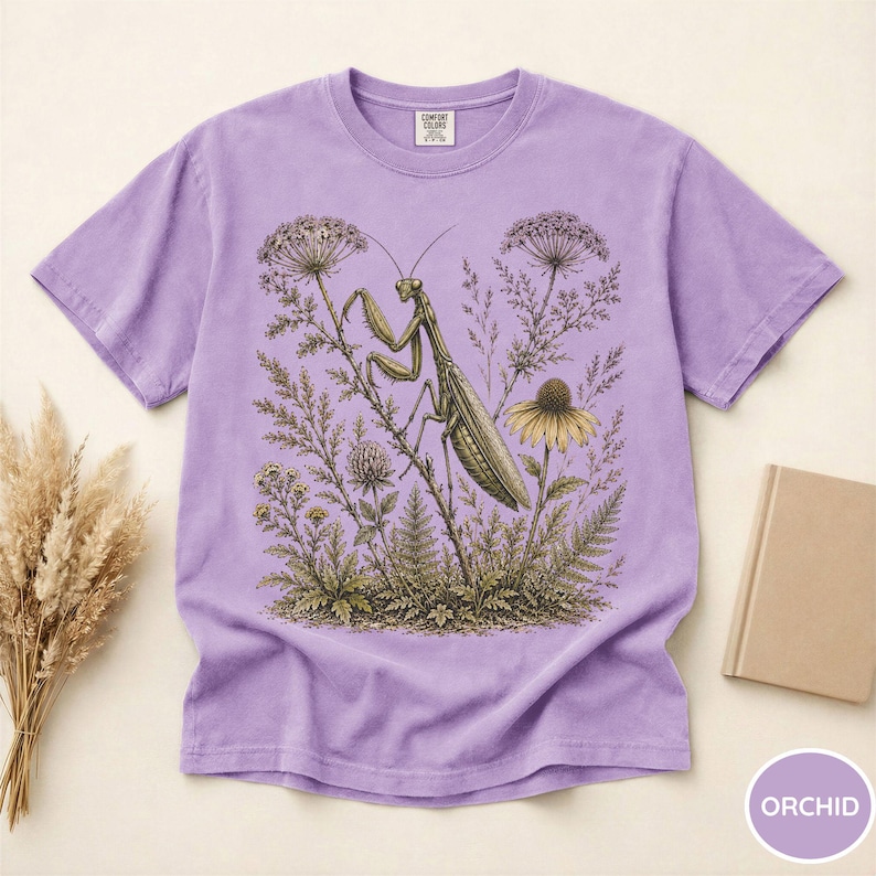May include: A light purple t-shirt featuring a detailed illustration of a praying mantis amidst wildflowers and foliage. The design is in shades of green, brown, and yellow, set against a soft purple background. The shirt has a classic crew neck and short sleeves.