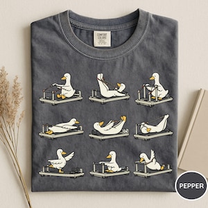 K&ouml;nnte beinhalten: Dunkelgraues T-Shirt mit einem Design von Cartoon-Enten, die auf Pilates-Reformern trainieren. Die Enten sind wei&szlig; mit gelben Schn&auml;beln und F&uuml;&szlig;en. Das T-Shirt hat einen Rundhalsausschnitt.