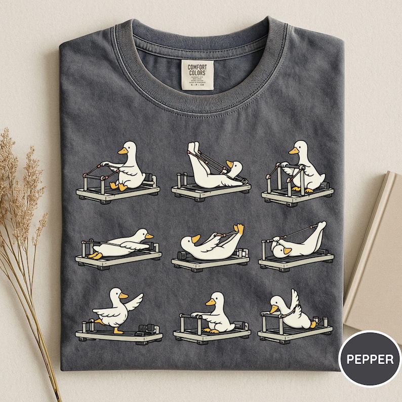 K&ouml;nnte beinhalten: Dunkelgraues T-Shirt mit einem Design von Cartoon-Enten, die auf Pilates-Reformern trainieren. Die Enten sind wei&szlig; mit gelben Schn&auml;beln und F&uuml;&szlig;en. Das T-Shirt hat einen Rundhalsausschnitt.