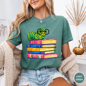 Pu&ograve; includere: Una t-shirt a maniche corte color verde salvia con una stampa grafica di un bruco verde brillante che indossa occhiali tondi neri. Il bruco &egrave; posizionato sopra una pila di cinque libri colorati nelle tonalit&agrave; del rosa, blu e giallo.