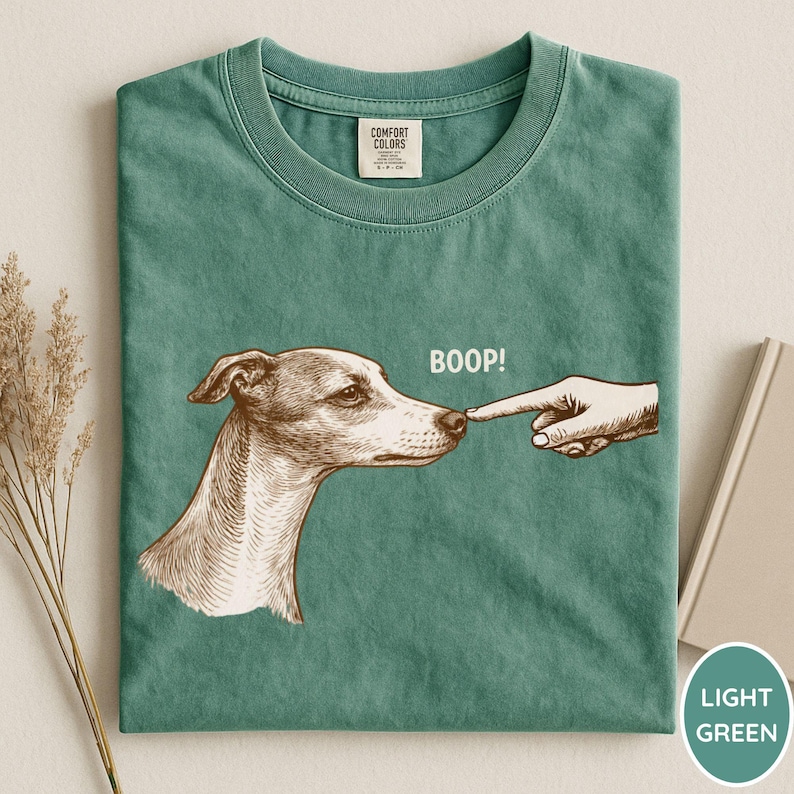 May include: A sage green t-shirt featuring a brown line-art illustration of a dog's head being touched on the nose by a human finger. The text "BOOP!" is printed in white above the dog's nose. The shirt has a Comfort Colors brand neck label.