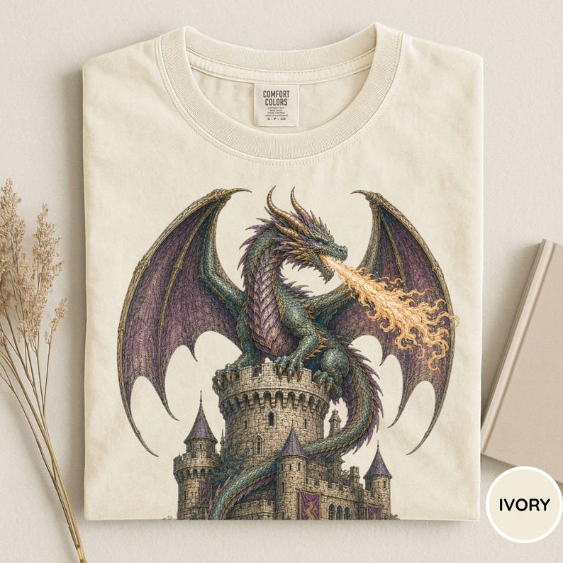May include: An ivory t-shirt featuring a graphic print of a green and purple dragon perched atop a stone castle tower, breathing a stream of orange fire. The shirt has a crew neckline and a Comfort Colors brand label printed on the inside collar.