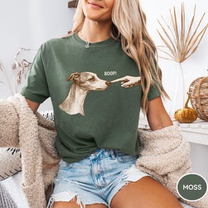 May include: A moss green short-sleeved t-shirt featuring a graphic illustration of a dog being touched on the nose by a human finger. The text BOOP! is printed in white above the dog's head. The shirt has a crew neckline and a relaxed fit.